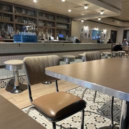 Super cute - The image shows an indoor restaurant setting with a bar in the background, stocked with glassware and bottles. The seating area includes a table with vintage-style chairs that have patterned upholstery and chrome legs. The floor has a black-and-white geometric tile pattern, and the ceiling has recessed lighting. There are a few patrons visible in the background.