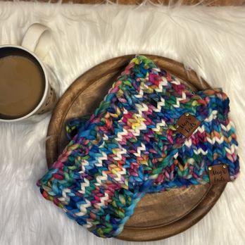 Fiber Fever - Custom headbands made with yarn from Fiber Fever - A multicolored, chunky knit blanket with tags labeled 'Meg's Knits' is placed on a wooden tray, next to a cup of coffee on a white furry surface. - Knitting Supplies Near Me - Milwaukee, Wisconsin