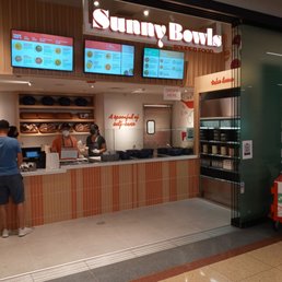Sunny Bowls soup counter located in a shopping mall food court, with three digital menu screens displaying soup options. The counter has staff wearing masks and aprons, with a customer placing an order. The decor includes wooden paneling and shelves with bowls and containers. A sign indicates self-care, and a QR code is visible on the side. An orange soup cart is positioned nearby.