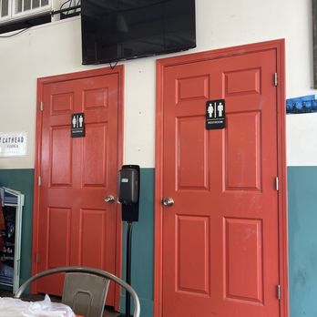The Station - Two red-painted doors with black restroom signs indicating male and female restrooms are positioned side by side on a white wall. A mounted flat-screen TV is installed above the doors. A hand sanitizer dispenser is located between the doors, and part of a table with chairs is visible in the foreground. The setting appears to be inside a casual dining or cafe environment. - Pizza Near Me - Jackson, Mississippi
