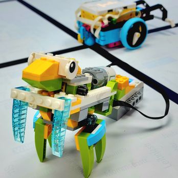 Zebra Robotics - Praying Mantis - Two LEGO robotic models are shown on a table with black lines. The foreground features a crab-like robot with green legs, a white and yellow body, and translucent blue claws. In the background, there is a wheeled robot with a white, blue, and black color scheme, connected by wires. The models are assembled with various LEGO pieces, and the table surface has black lines, possibly for robotics or coding activities. - Summer Camps Near Me - Toronto, Ontario
