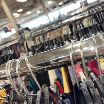 Value Outlets - A clothing rack with assorted garments hangs in a retail store, with other racks and shoppers visible in the background. - Men's Clothing Near Me - Farmingdale, New York