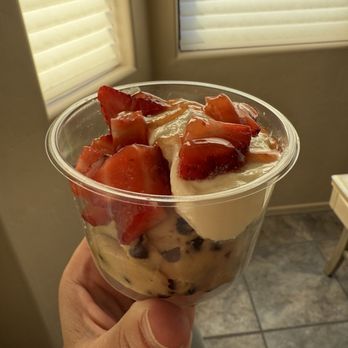 Cinnaholic - Cookie dough - A person holds a clear plastic cup filled with a layered dessert that includes sliced strawberries, cream, and chocolate chips. The background shows a window with closed blinds and a tiled floor, indicating an indoor setting. The dessert appears fresh and creamy, with visible fruit and chocolate elements. - Bakeries Near Me - Tucson, Arizona