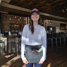 A woman with long brown hair, wearing a white long-sleeve shirt, a baseball cap, and a black apron with pens and notebooks in the front pocket, stands inside a bar or restaurant. The background features a bar counter with bottles, a brick wall, and hanging lights, indicating an indoor dining or drinking establishment.