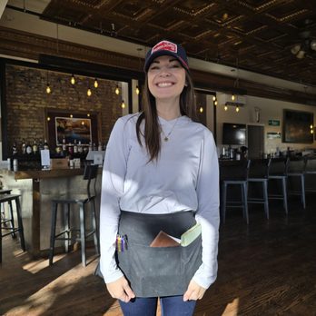 Ray's Corner Cafe - A woman with long brown hair, wearing a white long-sleeve shirt, a baseball cap, and a black apron with pens and notebooks in the front pocket, stands inside a bar or restaurant. The background features a bar counter with bottles, a brick wall, and hanging lights, indicating an indoor dining or drinking establishment. - Salad Near Me - Montrose, Colorado