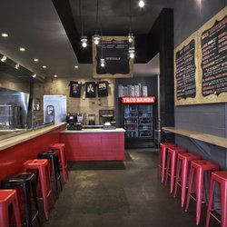 Taco Bamba Falls Church - Interior of restaurant with kitchen on the left with a glass divider and a small counter with stools. another small counter a...