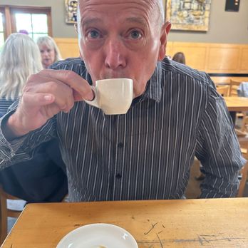 Downtown Subscription - My husband with his espresso and extra.....nope just EXTRA! - A man with a bald head and striped shirt is drinking from a small white coffee cup at a cafe, with a few women in the background. - Coffee & Tea Near Me - Santa Fe, New Mexico