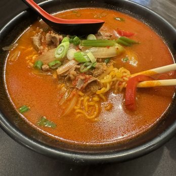 Yoodon - A black bowl contains spicy ramen with a red broth, noodles, sliced vegetables, and pieces of meat. Green onions are sprinkled on top. A red spoon rests on the edge of the bowl, and chopsticks are placed inside, holding some noodles and a slice of red pepper. The bowl is on a dark table, with a small potted plant and a person’s legs visible in the background. - Korean Near Me - Bayside, New York