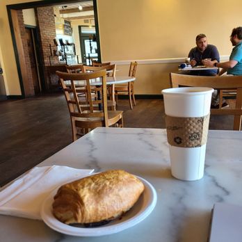 Athan's Bakery - Chocolate croissant! - A croissant on a small white plate and a disposable coffee cup with a cardboard sleeve are on a marble table in a cafe. The cafe has wooden chairs, a yellow wall with lighting, and a doorway leading to another area with brick walls and shelves. Two men are seated at a table in the background, engaged in conversation. - Bakeries Near Me - Brookline, Massachusetts