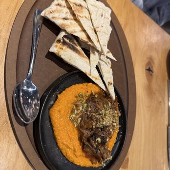 Kitchen Social - Short rib and carrot hummus - The image shows a black oval plate containing a serving of orange-colored hummus topped with shredded short rib, chopped green onions, and crushed nuts. The hummus is accompanied by several pieces of grilled flatbread, arranged on a brown oval tray. A metal spoon rests on the tray, and the setting appears to be on a wooden table, likely in a restaurant environment. - Cocktail Bars Near Me - Columbus, Ohio