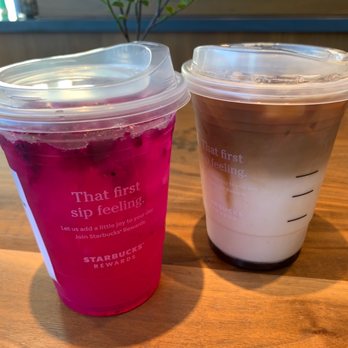 Starbucks - Mango dragonfruit iced tea & Iced Apple Crisp macchiato - The image shows two Starbucks iced beverages on a wooden table. The drink on the left is bright pink with visible berries, while the drink on the right has a layered appearance with white and brown sections, likely a coffee or tea-based drink. Both cups have clear plastic lids and Starbucks branding, with the pink drink featuring the phrase 'That first sip feeling.' in white text. - Coffee & Tea Near Me - Exton, Pennsylvania
