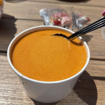 Champlin's Seafood - Lobster Bisque - A white disposable cup contains lobster bisque, a creamy orange seafood soup, with a spoon inserted. The cup is placed on a wooden table, with a shadow cast on the surface. In the background, there are other food items, including a plate of fried seafood and a glass of beer, along with condiment packets and small containers. The setting appears to be a casual seafood restaurant or outdoor dining area. - Seafood Near Me - Narragansett, Rhode Island