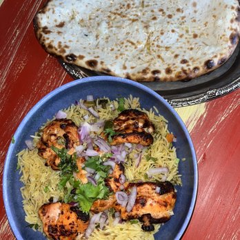 Chit Chaat - Chicken biryani tandoori style & Garlic Cheese Naan - The image shows a meal consisting of grilled tandoori chicken pieces garnished with chopped onions and cilantro, served on a bed of yellow rice. A large piece of garlic cheese naan is placed on a silver tray above the plate. The meal is presented on a red wooden table, typical of Indian or Pakistani cuisine settings. - Indian Near Me - Racine, Wisconsin
