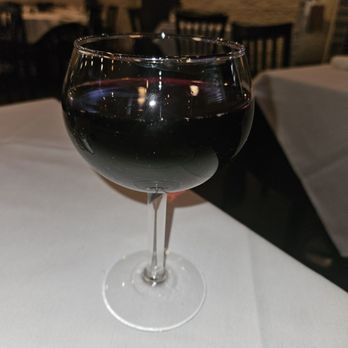 Casa Bella - A clear wine glass filled with red wine is positioned on a white tablecloth in a restaurant. The background features dark wooden chairs and tables, with soft lighting creating a cozy atmosphere. The image captures the glass and its contents with reflections on the surface of the wine. - Italian Near Me - Scranton, Pennsylvania