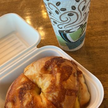 Mt. Taylor Coffee Co - Loaded Bagel Sandwich and Carmel Latte - A paper coffee cup with a plastic lid and decorative sleeve featuring coffee beans and a green design is on a wooden table. Next to it, a white takeout container holds a pastry with a golden-brown, slightly flaky crust. The setting appears to be a café or coffee shop. - Coffee & Tea Near Me - Grants, New Mexico