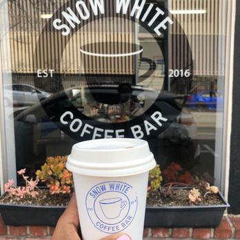 Snow White Coffee - My hand holding a Chai latte in white cup with a blue stamp of the company logo in front of the shop window. - Coffee & Tea Near Me - Oakland, California Snow White Coffee - My hand holding a Chai latte in white cup with a blue stamp of the company logo in front of the shop window. - Coffee & Tea Near Me - Oakland, California