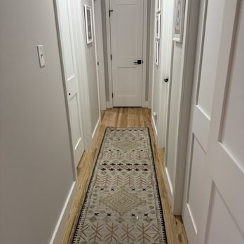 Denver Remodels - Doors/base/case - A hallway with white walls and multiple doors, decorated with framed pictures, and a patterned rug on wooden floor. - General Contractors Near Me