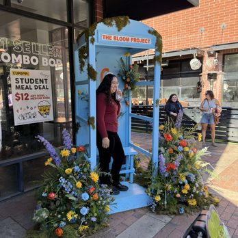 Tempe Blooms - A woman with glasses and dark hair, wearing a maroon top and black pants, stands inside a blue floral booth labeled 'The Bloom Project' on a sidewalk. The booth is decorated with colorful flowers and greenery. In the background, there are pedestrians, a brick building, and a storefront with signs advertising a student meal deal. The scene is outdoors with clear weather. - Festivals Near Me - Tempe, Arizona