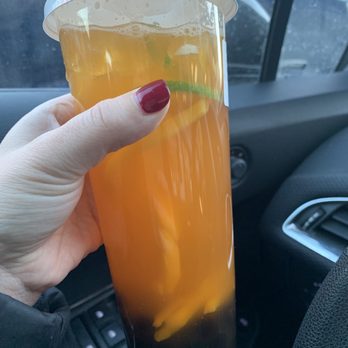 Royal Tea - Mango Passion tea - A person with red nail polish holds a tall plastic cup of mango passion tea with lemon slices, inside a vehicle. - Bubble Tea Near Me - Waltham, Massachusetts