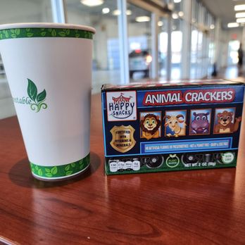 Fiesta Subaru - Coffe and snack in the service area! Bright and cheerful. - A paper cup with a green leaf pattern and a box of Happy Snacks animal crackers are positioned on a wooden table. The background shows a store interior with glass windows, doors, and a few people. The setting appears to be a retail or service area, possibly within a dealership or similar establishment. - Car Dealers Near Me - Albuquerque, New Mexico