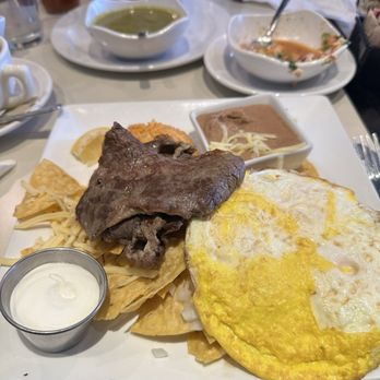 Cafe con Leche - Chaq Chilaquiles Verdes - A breakfast plate featuring eggs, beef, cheese, and sides, with bowls of salsa and sauces in the background at Cafe con Leche, Chicago. - Mexican Near Me - Chicago, Illinois