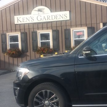 Ken's Gardens - The image shows the exterior of Ken's Gardens, a garden center with a wooden facade, a sign displaying the business name, and windows with dark shutters and flower boxes. A black vehicle is parked in front of the building, partially obscuring the view of the storefront. An illuminated 'Open' sign is visible in one window, and the sky appears partly cloudy with some trees in the background. - Nurseries & Gardening Near Me - Lancaster, Pennsylvania