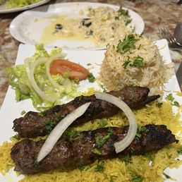 Beef kebab platter, kofta kebab? So good! - A plate features grilled meat skewers on yellow rice, garnished with onions and herbs, alongside a rice mound and a small salad. A creamy dish is visible in the background.