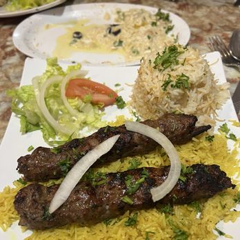 Hala Cafe - Beef kebab platter, kofta kebab? So good! - A plate features grilled meat skewers on yellow rice, garnished with onions and herbs, alongside a rice mound and a small salad. A creamy dish is visible in the background. - Mediterranean Near Me - Euless, Texas