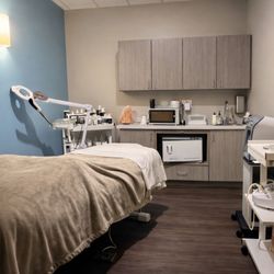Hand & Stone Massage and Facial Spa - Professional facial and massage treatment room with plush treatment bed, magnifying lamp, skincare equipment, Himalayan salt ...