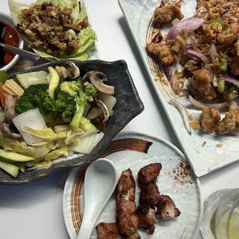 Juicy Dumplings - 2 Chicken Lettuce Wrap Salt Pepper Style chicken  Chicken skewers - The image shows a table with multiple Asian dishes. A small plate contains fried chicken pieces with a crispy exterior. A larger rectangular plate holds stir-fried vegetables, including broccoli, mushrooms, and cabbage. Another dish features a minced meat salad served on a lettuce leaf, garnished with onions and chili peppers. The dishes are presented on different plates and bowls, with a white table surface visible. - Chinese Near Me - Euless, Texas