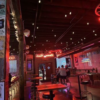 Broken Hearts - A bar interior featuring brick walls, tables, bar stools, and neon signs, with some patrons seated and standing, illuminated by string lights and ambient lighting. - Wine Bars Near Me - Chicago, Illinois