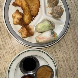 AP1. Ap Combo - A round, divided plate contains Thai appetizers such as fried fish or chicken skewers, fried wontons, steamed dumplings, and fresh spring rolls with visible vegetables and shrimp. The plate is placed on a wooden table. Below, a smaller plate holds three small containers of dipping sauces, including soy sauce and peanut sauce. The presentation suggests a shared appetizer platter at a Thai restaurant.