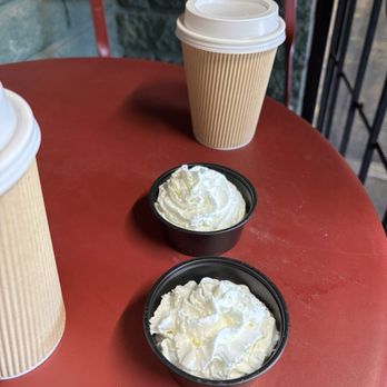 Greer Grounds - Pup cups - The image shows two black plastic cups filled with whipped cream placed on a red table. A brown paper coffee cup with a white plastic lid is positioned behind the whipped cream containers. The setting appears to be a cafe or outdoor seating area, with a red chair and a stone wall visible in the background. The table surface has some minor marks or stains. - Coffee & Tea Near Me - Greer, Arizona