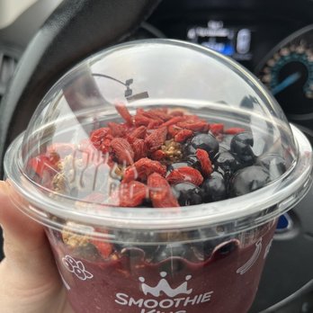 Smoothie King - a hand holding a cup of smoothie - A clear plastic cup with a domed lid contains a smoothie bowl topped with blueberries, goji berries, and granola. The cup features the 'Smoothie King' logo and is held by a person's hand inside a vehicle. The dashboard and steering wheel are visible behind the cup, indicating the photo was taken inside a car. - Juice Bars & Smoothies Near Me - Newburgh, New York