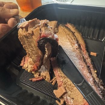 Truth BBQ - The bend on the brisket, almost fell apart. - A sliced smoked brisket with a dark bark and tender meat is placed on a black tray, with a glass of iced tea in the background. - Barbeque Near Me - Brenham, Texas