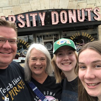 Varsity Donuts - Family selfie - A family of four poses for a selfie in front of University Donuts, a donut shop with a vintage sign, on a cloudy day. - Donuts Near Me - Manhattan, Kansas