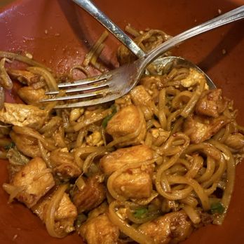 Dragon Express - Chicken Pad Thai - A red bowl contains chicken pad thai with stir-fried noodles, chicken pieces, and vegetables. A metal fork and a spoon are placed in the bowl, with some noodles and chicken visible. The dish appears to be partially eaten, with remnants of sauce and ingredients. The background shows a wooden table and part of a person's hand holding the bowl. - Chinese Near Me - Mesa, Arizona