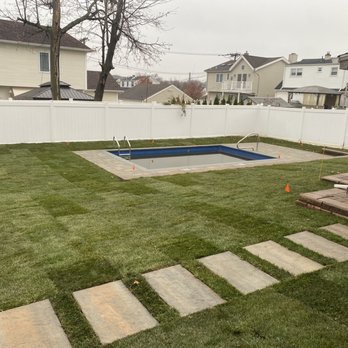 The Pool Boss - The image shows a backyard with a rectangular in-ground swimming pool, partially filled with water, surrounded by a paved stone border. The yard is enclosed by a white privacy fence, with neighboring houses and trees visible in the background. There is a small deck area with a scooter and some outdoor furniture on the right side. The grass is well-maintained, and the sky is overcast. - Pool & Hot Tub Service Near Me - Wayne, New Jersey