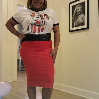 Sidecca - Work flow in my cute Santa vixen tshirt  from sidecca Pasadena - A woman with shoulder-length hair and a cheerful expression stands indoors, wearing a white graphic t-shirt with a cartoon design, a black belt, and a red fitted pencil skirt. She has a necklace with a pendant and black shoes with socks. The room features light-colored walls, a framed black-and-white photograph of a woman on the wall, and a doorway leading to another room. - Accessories Near Me - Altadena, California