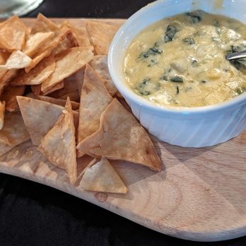 The Eis House - Spinach and Artichoke - A white ramekin filled with spinach and artichoke dip, containing visible spinach leaves and artichoke pieces in a creamy mixture, is placed on a wooden serving board. Next to the dip, there is a pile of triangular tortilla chips. The presentation suggests a shared appetizer at a restaurant or event setting. - American Near Me - Mexico, New York