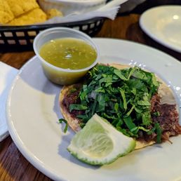 Barbacoa Taco - A taco topped with chopped fresh herbs, served with a lime wedge and a small cup of green salsa on a white plate. The background includes a basket of tortilla chips and a bowl, indicating a Mexican restaurant setting.