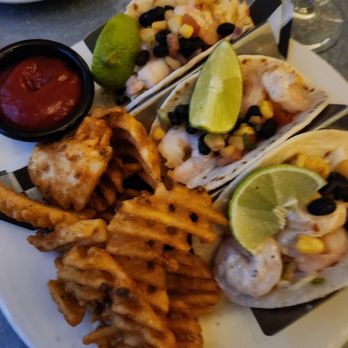 Iconic  - Shrimp tacos - A white plate holds three shrimp tacos garnished with lime wedges, corn, and black beans. The tacos are served with a side of waffle fries and a small container of salsa. The presentation suggests a seafood dish with Mexican-inspired toppings, suitable for casual dining. - Cocktail Bars Near Me - Utica, New York