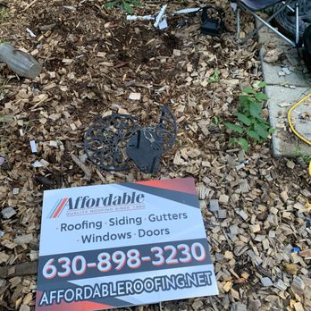 Affordable Roofing - The image shows a yard area covered with wood chips, featuring a fallen decorative metal piece, a sign for Affordable Roofing with contact information, and scattered debris including a plastic bottle and some broken objects. There are small plants growing among the wood chips, and a fence is visible in the background. The scene appears to be an outdoor space with signs of recent disturbance or cleanup. - General Contractors Near Me - Aurora, Illinois