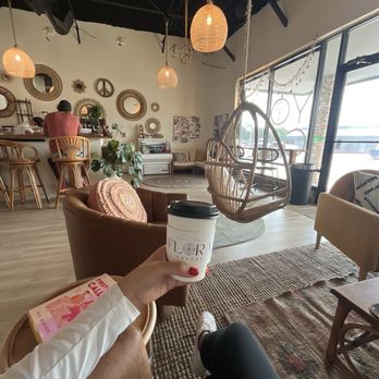 Flora Coffee - Inside Flora Coffee, a cafe with bohemian decor, hanging chairs, and warm lighting, a person holds a coffee cup with the cafe's logo. - Coffee & Tea Near Me - Austin, Texas