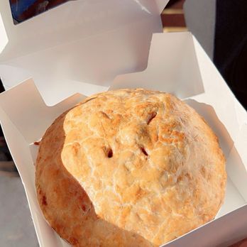 Jericho Cider Mill - Apple pie - A large apple pie with a golden, flaky crust is placed inside a white cardboard box. The pie appears freshly baked, with a slightly uneven surface and small slits on top. The person holding the box is outdoors, with sunlight illuminating the scene. The background shows a paved surface and part of a person's hand and arm. - Fruits & Veggies Near Me - Jericho, New York