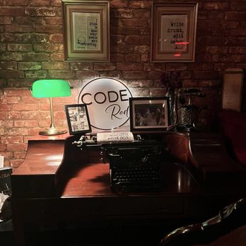 CODE RED - A vintage black typewriter is positioned on a polished wooden desk with a curved design, set against a brick wall. The desk features framed photographs, a green desk lamp, and decorative signs, including one that reads 'CODE Red.' Two framed quotes are hung on the brick wall above the desk. The scene has a warm, dimly lit ambiance, evoking a nostalgic or retro atmosphere. - Cocktail Bars Near Me - Washington, DC, District of Columbia