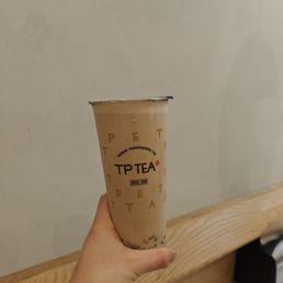 A hand is holding a cup of bubble tea from TP TEA, featuring tapioca pearls at the bottom. The cup has branding with the words 'TP TEA' and 'Taiwan Professional Tea,' and is set against a plain, light-colored wall with a wooden surface below. The image focuses on the beverage and hand, with no other objects or people visible.