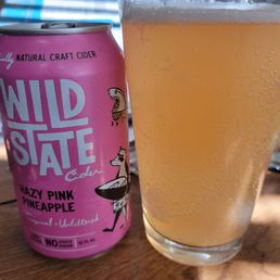 Hazy Pink Pineapple - A pink can of Wild State Cider Hazy Pink Pineapple is placed beside a glass filled with a light, bubbly cider, on a wooden surface.