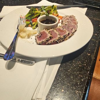 BrickTop's - Au tuna steak - A white plate holds slices of seared tuna with a pink center, coated with sesame seeds. Accompanying the tuna are mixed vegetables including broccoli, carrots, and snap peas, along with a small bowl of dark dipping sauce. The plate is set on a black granite bar counter with a fork, napkin, and utensils nearby. The background shows bottles of alcohol and bar equipment, indicating a restaurant or bar setting. - New American Near Me - Frontenac, Missouri
