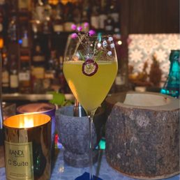 Diamond fizz with fresh organic basil - A cocktail with a yellow-green hue is served in a tall, elegant glass, garnished with a lemon slice, a cherry, and small purple flowers. The drink is placed on a blue napkin on a bar counter, surrounded by candles, a large wooden bowl, and decorative containers. The background features a blurred bar shelf with bottles and warm lighting.